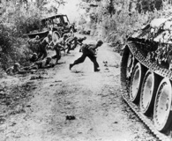 July 1944. American infantrymen dash across a hedgerow-bordered lane near St. Lo. A knocked-out German Panther tank sits in the foreground. National Archives.